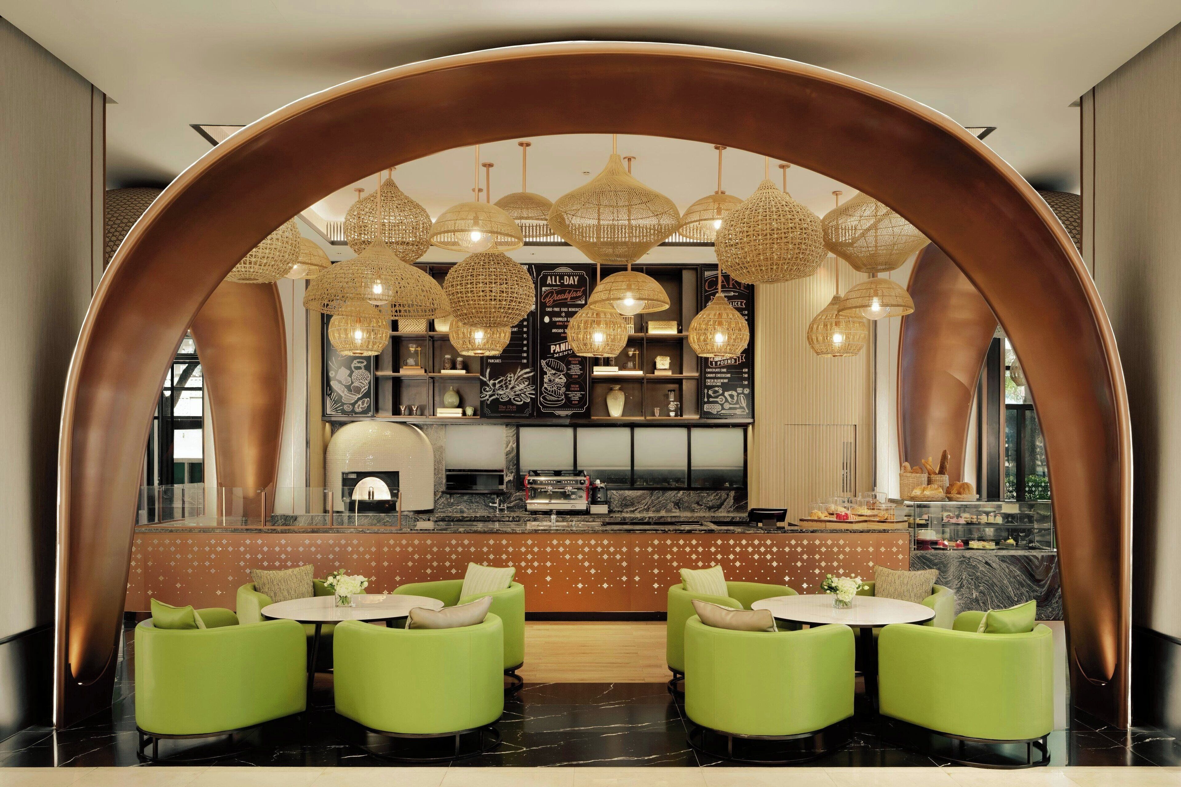 Un restaurant moderne avec des sièges verts, un bar central et des lustres en osier suspendus au plafond.