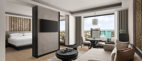 A modern hotel room with a bed, a sofa, a dining area, and a large window offering a city view.