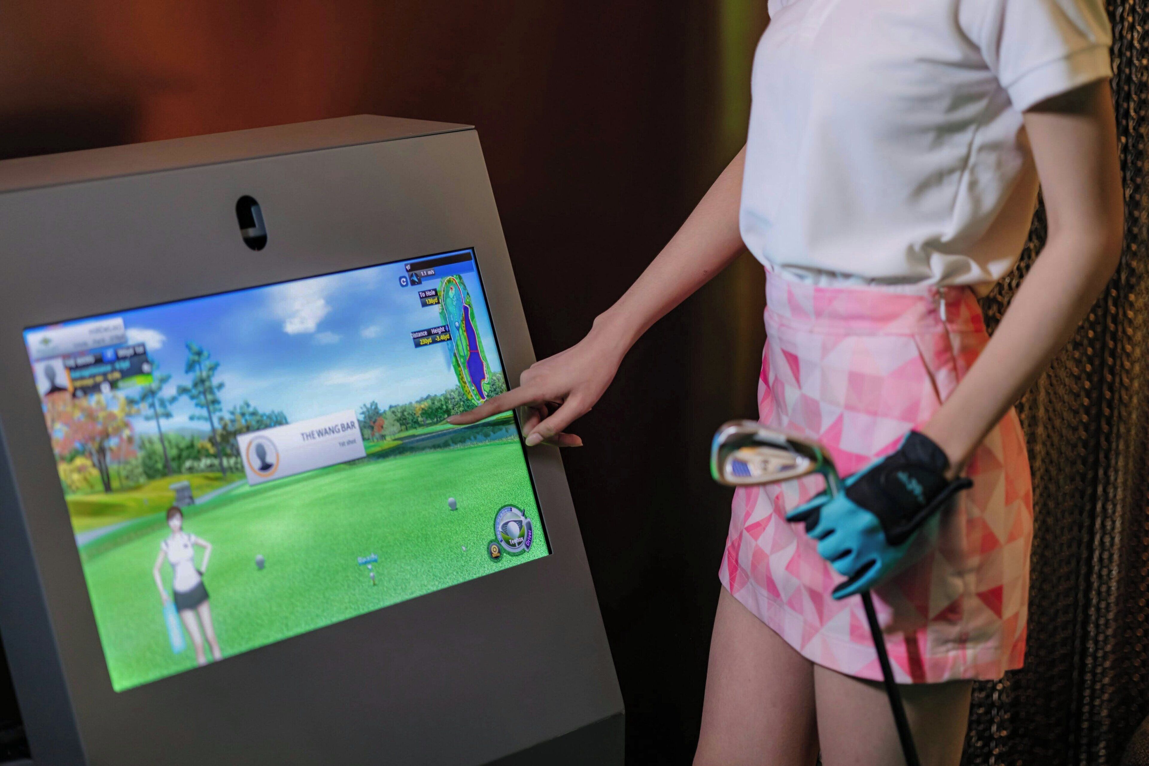 A person interacting with a large touchscreen display, possibly a golf simulation game, with a grassy field and a character on the screen.