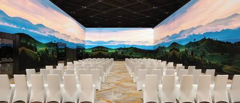 Un salón de conferencias con filas de sillas blancas, una alfombra estampada y un mural grande de un paisaje en las paredes.