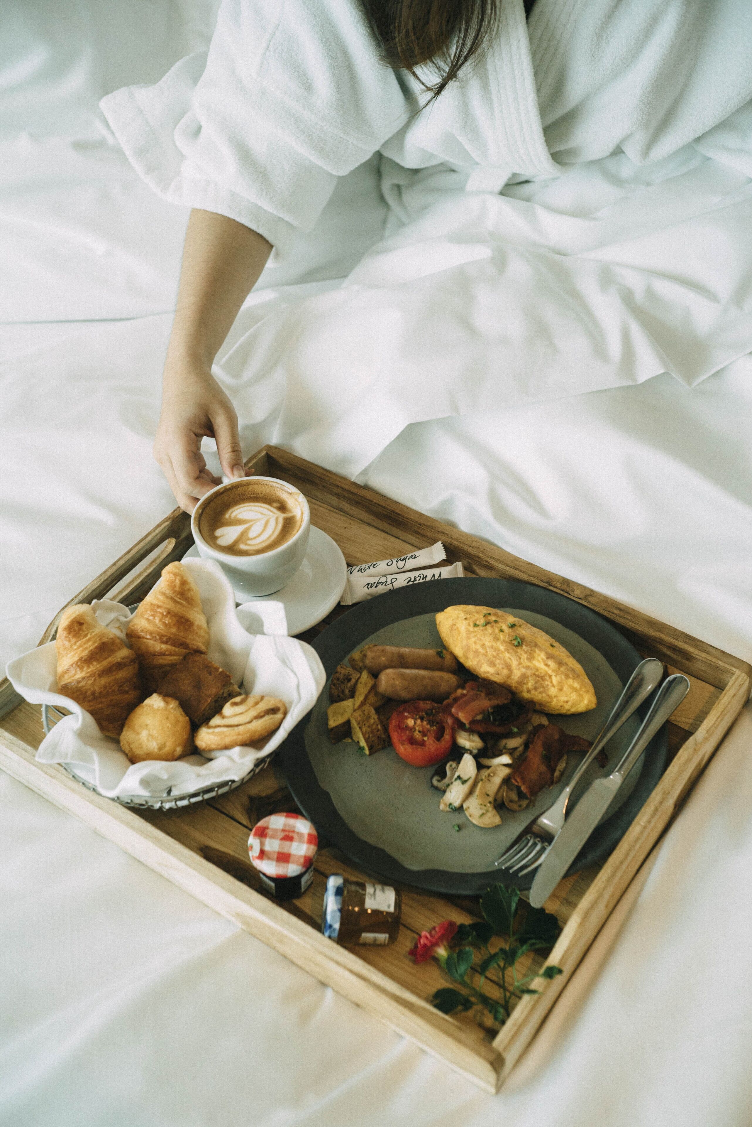 Een dienblad met ontbijtartikelen, waaronder croissants, eieren, tomaten en champignons, vergezeld van een kop koffie en een vork en mes op een bed.