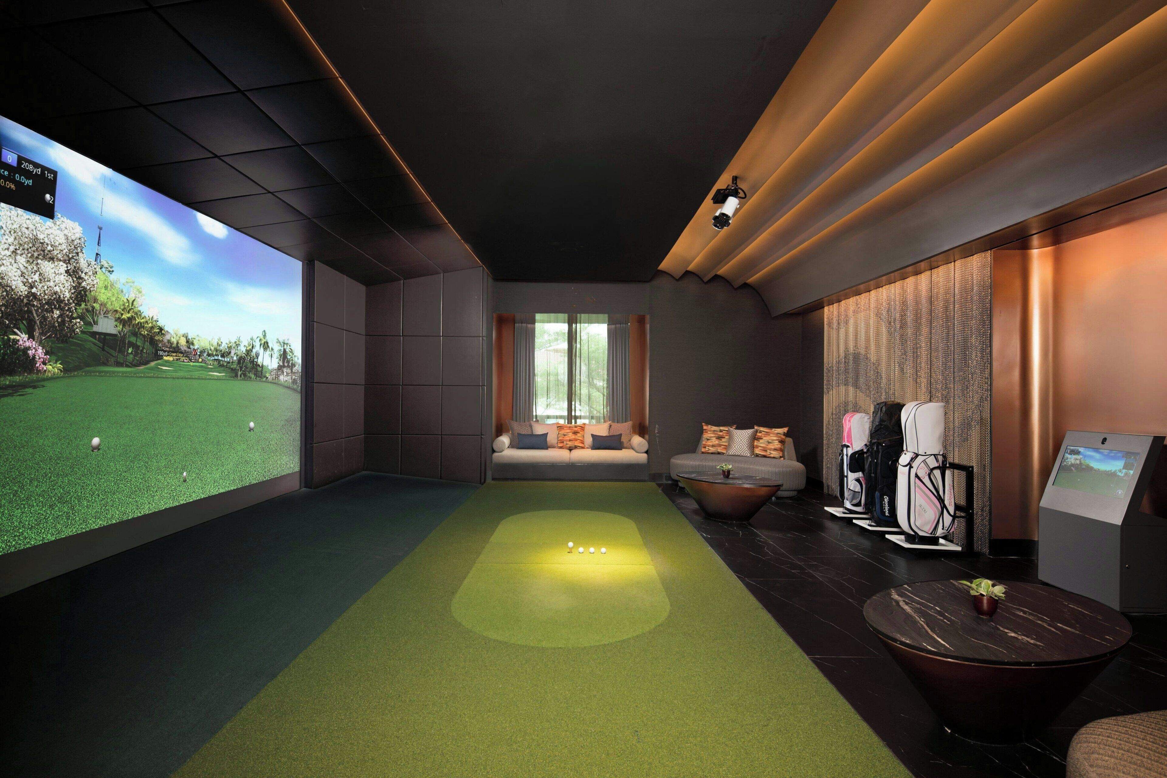 A room with a large screen displaying a golf course, a green carpeted area, a sofa with cushions, and a seating area with a view of trees.