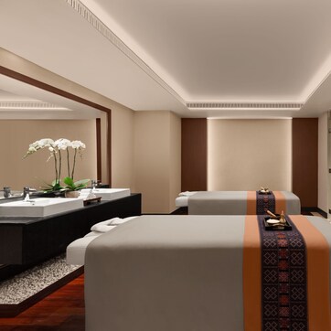 A modern hotel room with a large bed, a separate sofa, a glass-enclosed shower, and a vanity with two sinks.