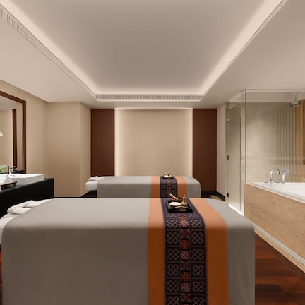 A modern hotel room with a large bed, a separate sofa, a glass-enclosed shower, and a vanity with two sinks.