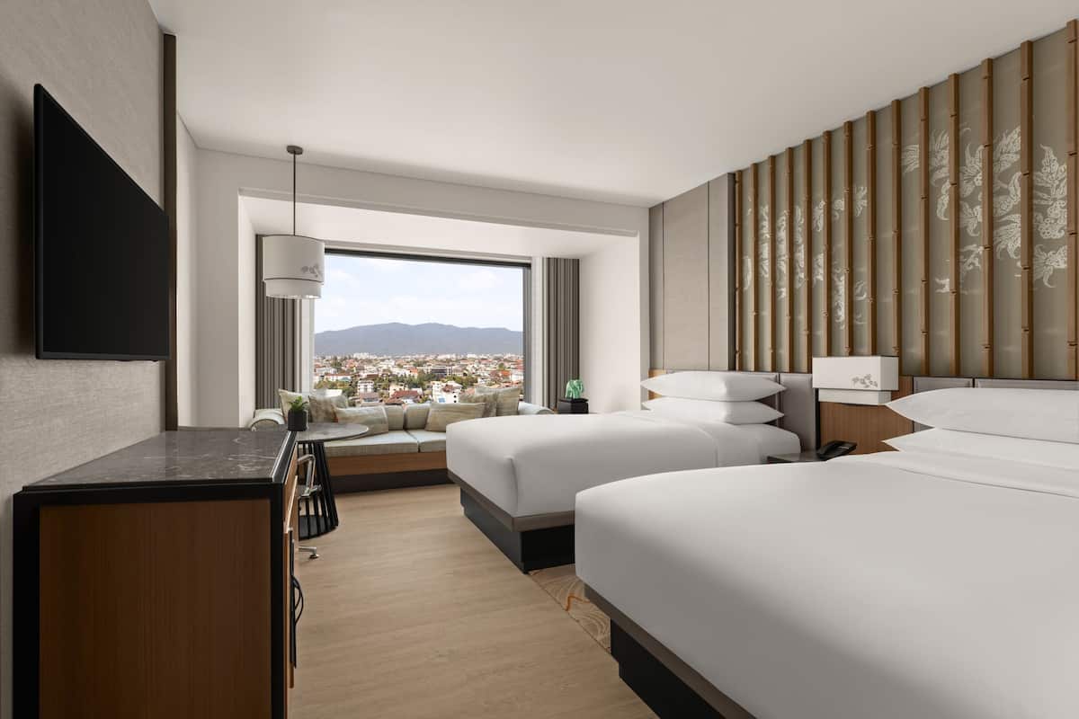 A modern hotel room with a large window offering a city view, a flat-screen TV, a desk, and two beds with a decorative headboard.