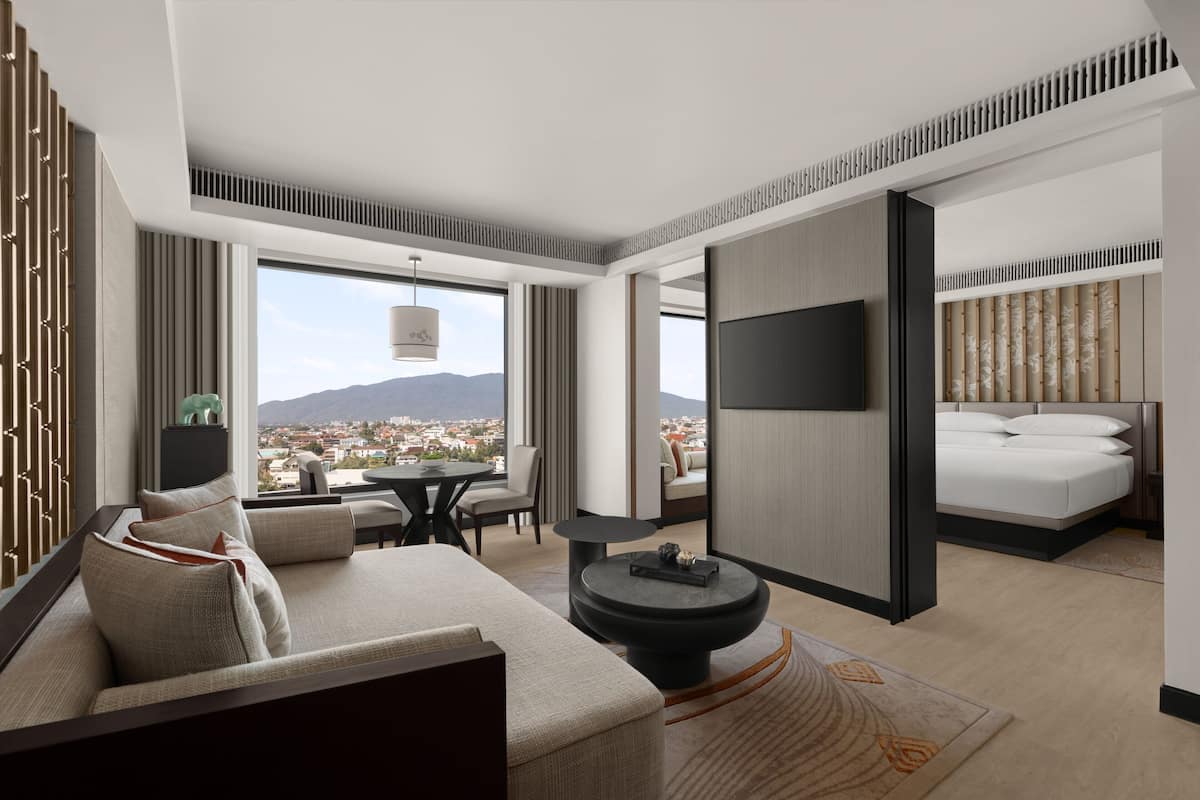 A modern hotel room with a sofa, a round coffee table, a bed, and a TV. The room has large windows offering a city view.