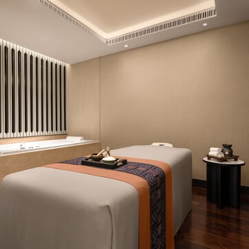 A modern spa room with two massage tables, a large mirror, a wooden reception desk, and a small side table with a plant.