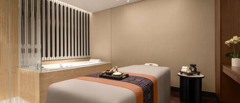 A modern spa room with two massage tables, a large mirror, a wooden reception desk, and a small side table with a plant.