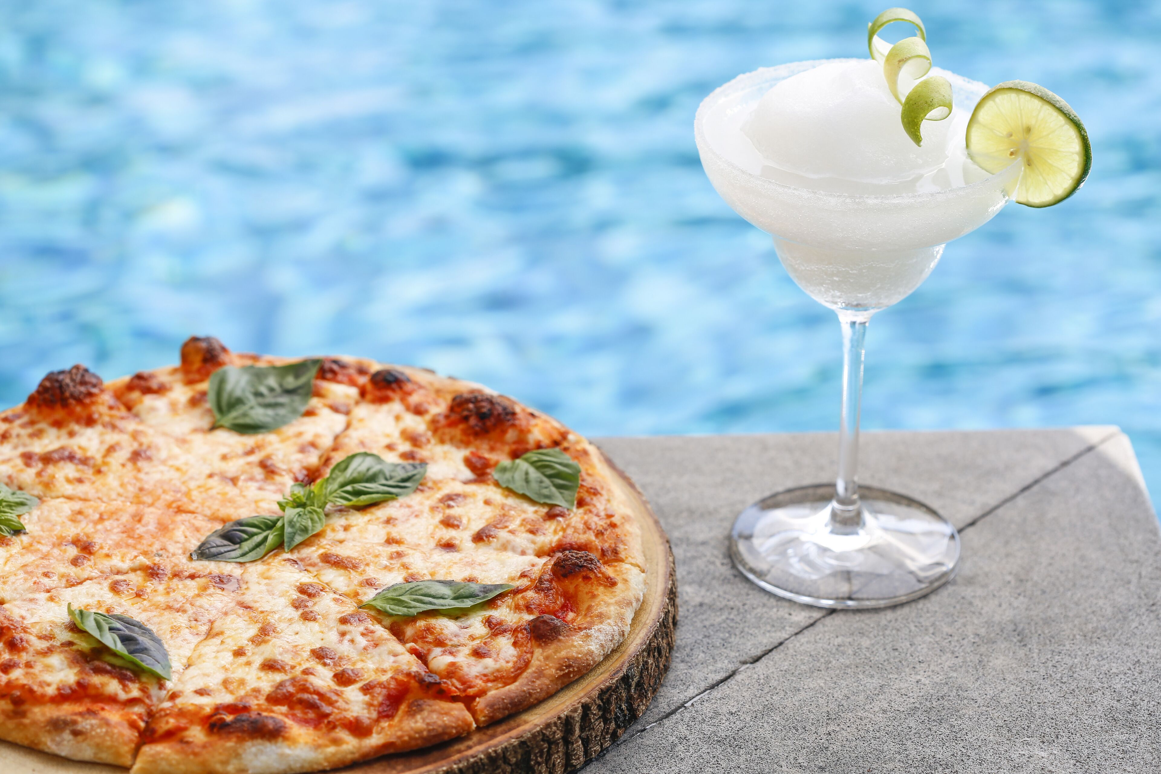 A pizza and a cocktail by a poolside.