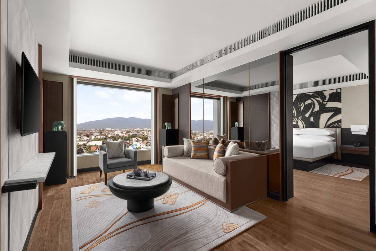 A modern hotel room with a sofa, armchair, and a dining table. There is a large window offering a city view.