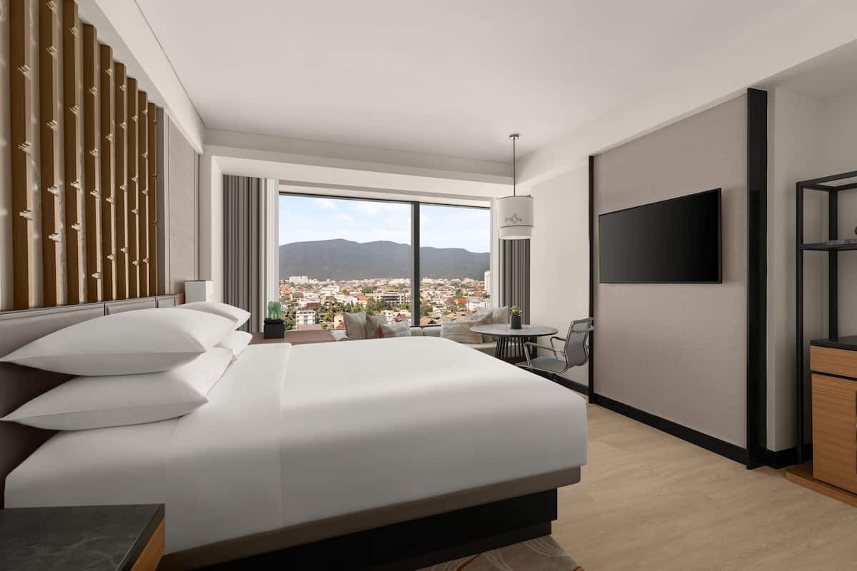 A modern hotel room with a large bed, a TV, a desk, and a view of a cityscape through large windows.