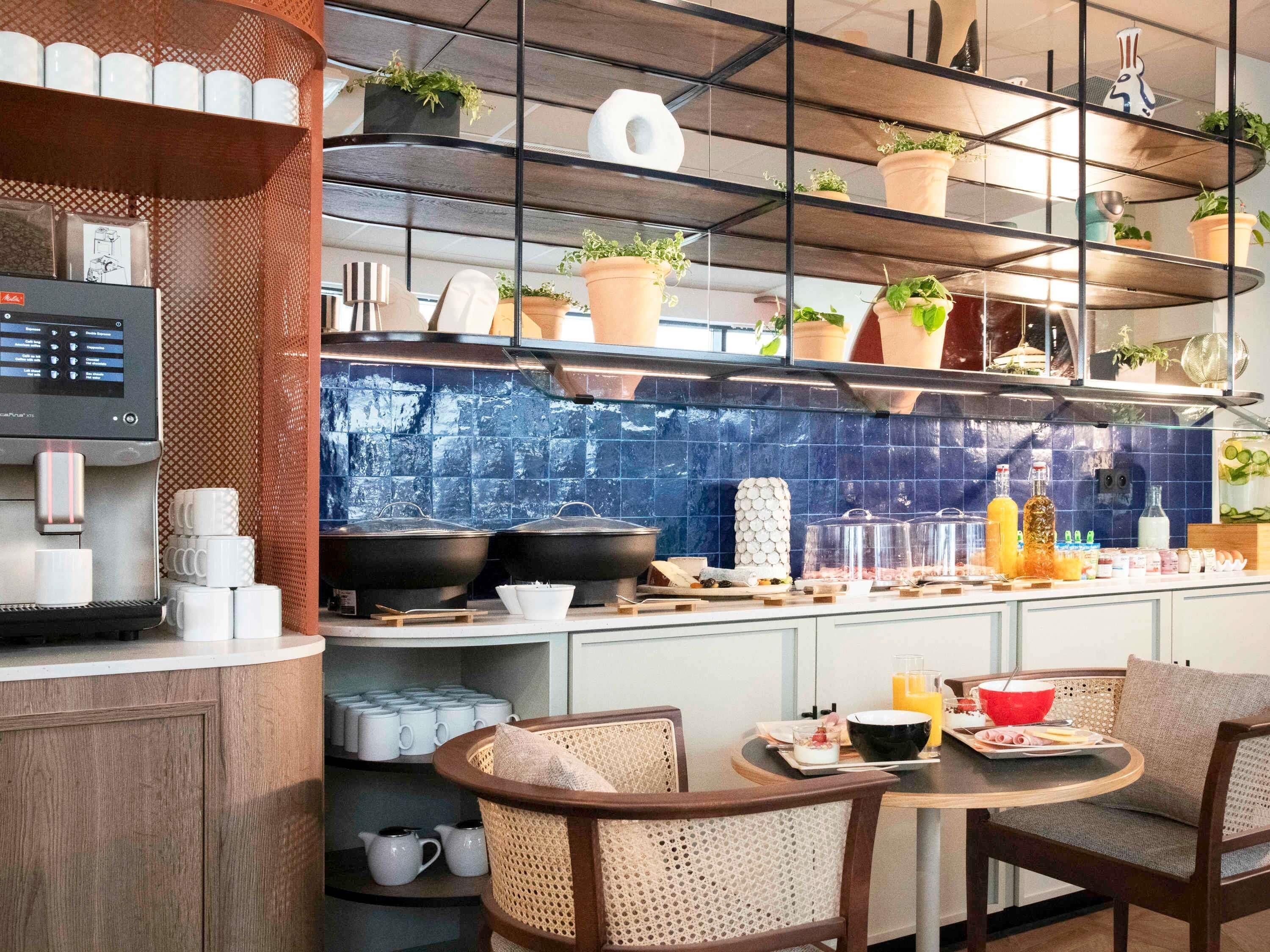A modern kitchen with a coffee machine, shelves with plants, and a dining area with chairs and a table.