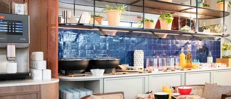 A modern kitchen with a coffee machine, shelves with plants, and a dining area with chairs and a table.