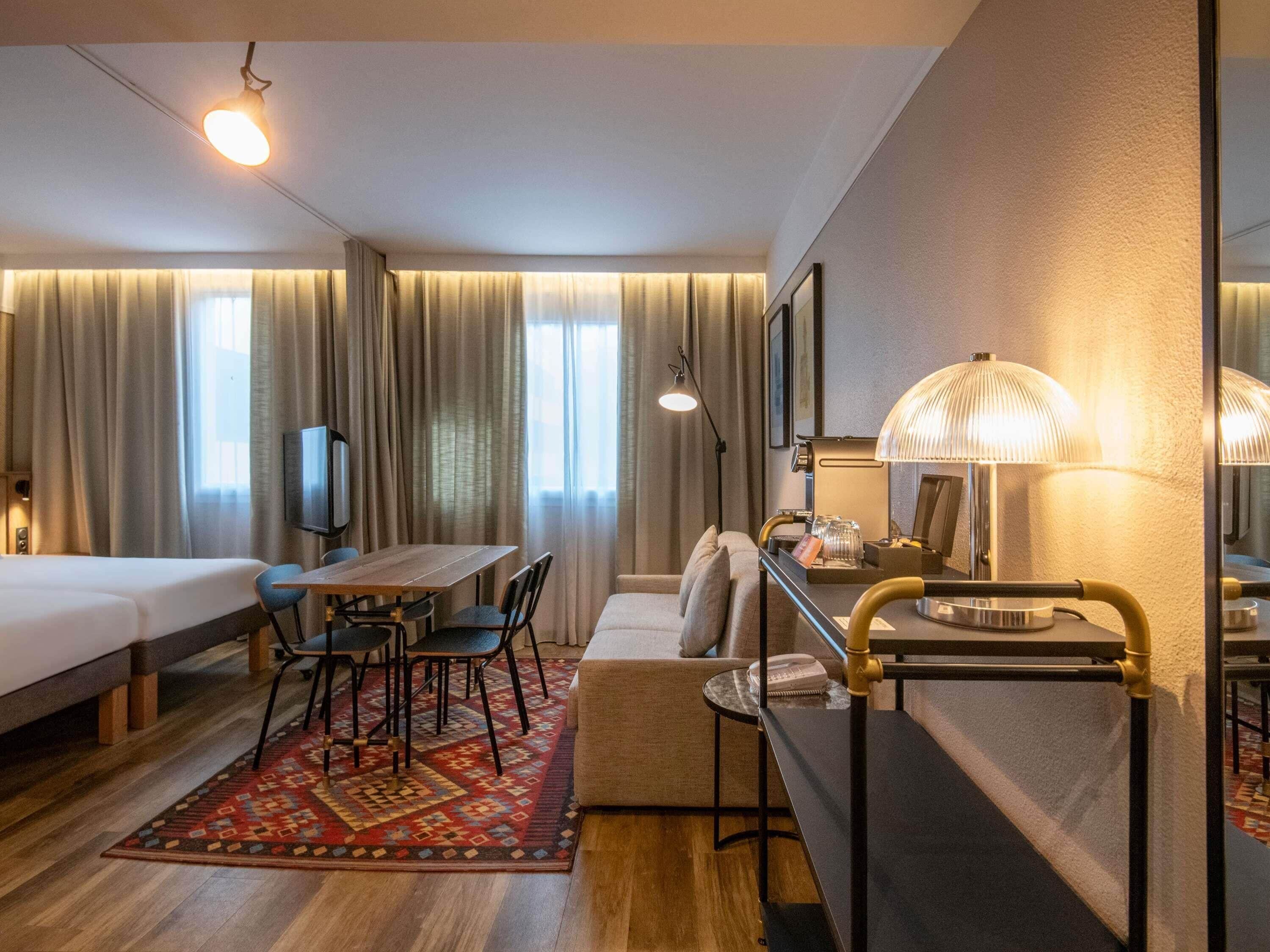 A hotel room with a bed, a desk, a chair, a sofa, a TV, a lamp, and a window with curtains.