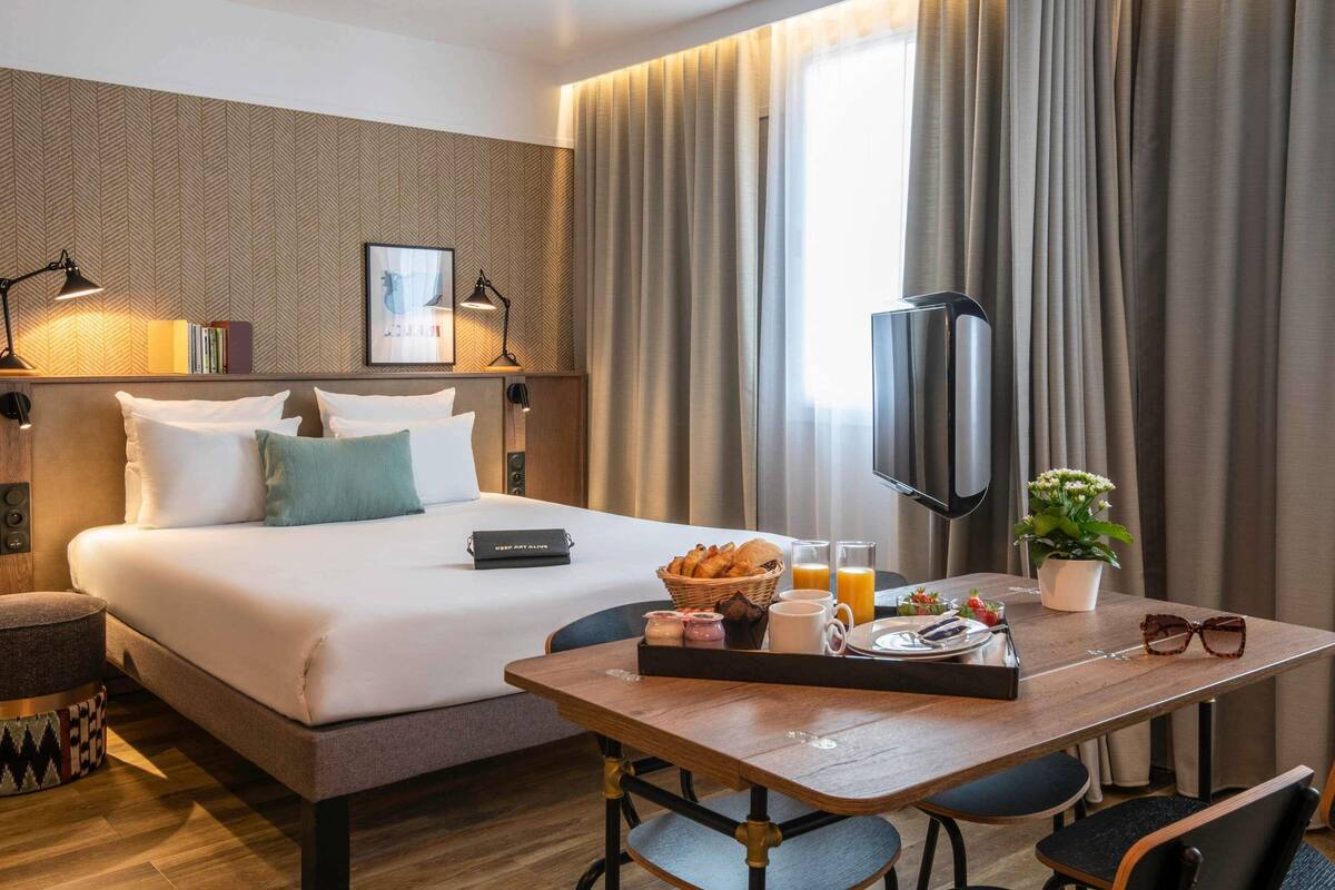 A hotel room with a bed, bedside lamps, a TV, a dining table with breakfast, and a potted plant.