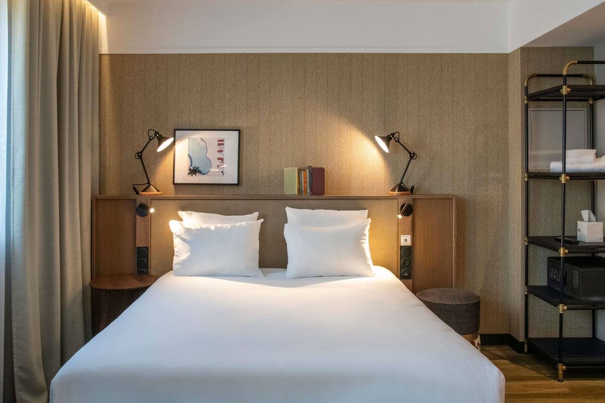 A hotel room with a large bed, two bedside tables, a headboard with a framed picture, a shelf with books and a lamp, and a wardrobe with a mirror.