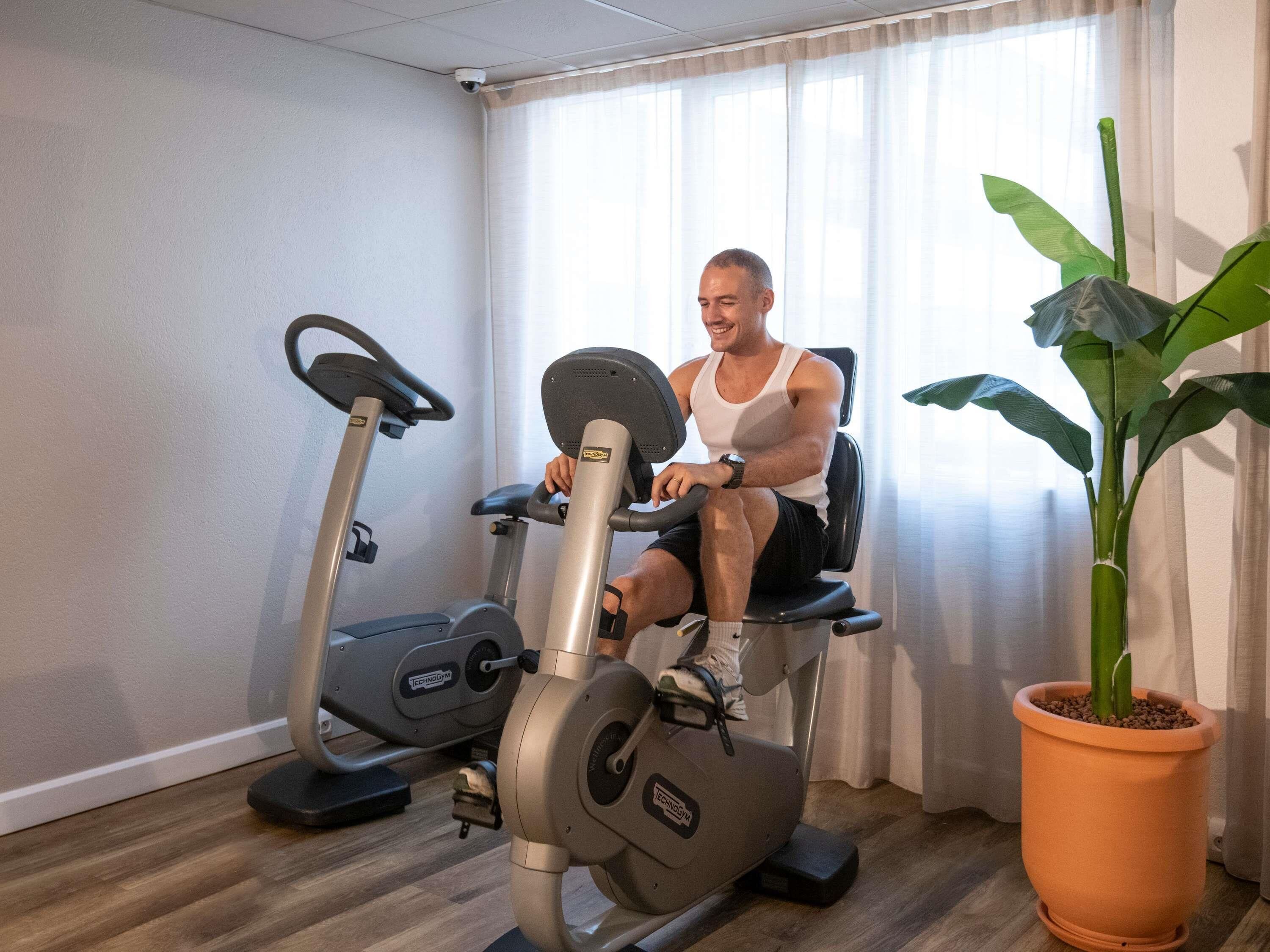 A man exercising on a stationary bike in a room with a potted plant and an exercise machine.