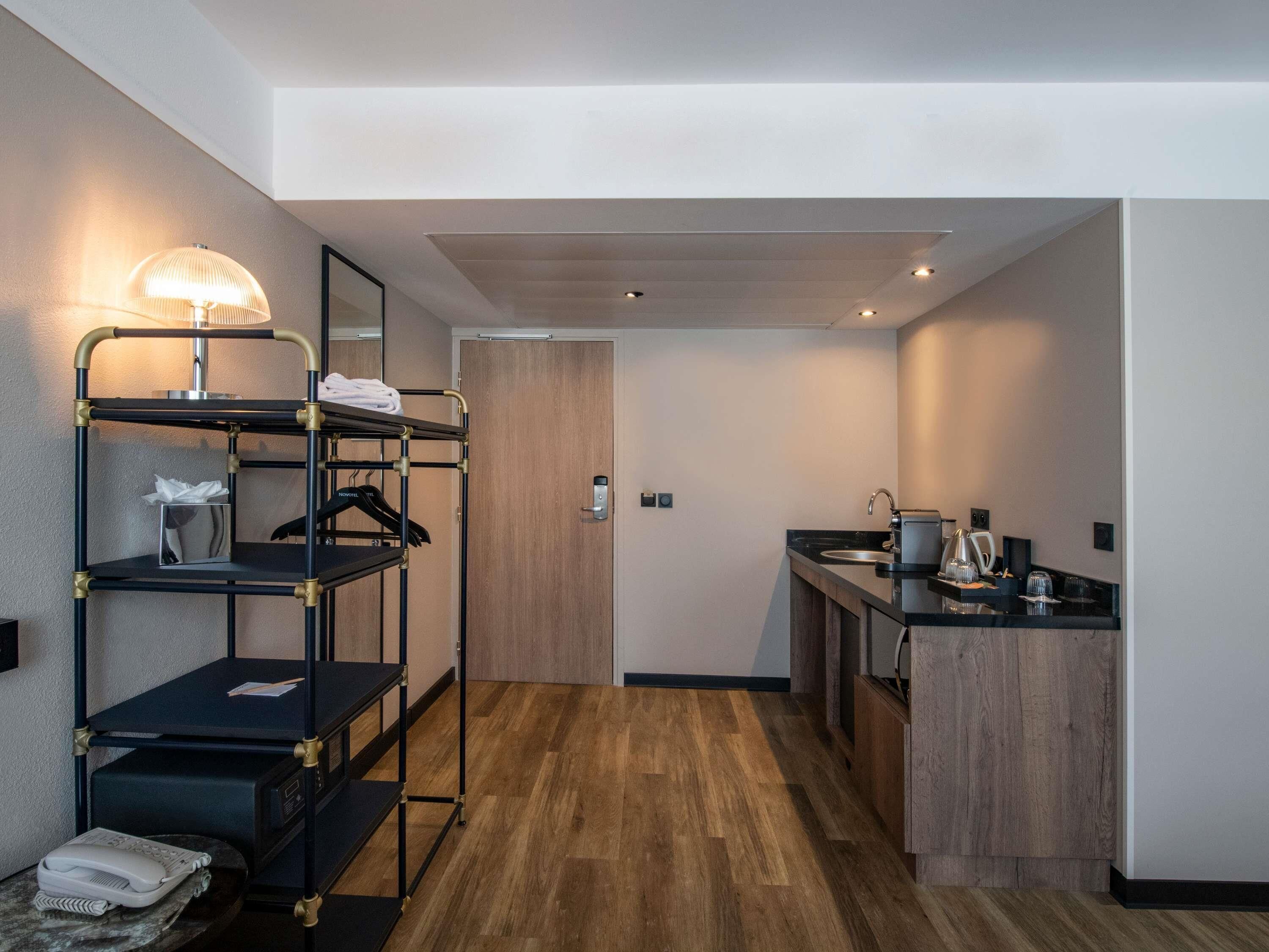 Quarto de hotel compacto com beliche, pequena cozinha e piso de madeira, e porta que dá para outro espaço.