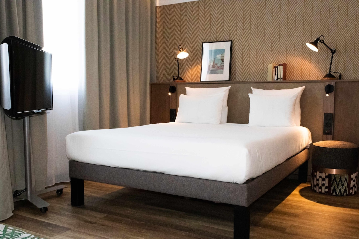 Una habitación de hotel moderna con una cama grande, un televisor de pantalla plana, una mesita de noche con lámpara y un cuadro enmarcado en la pared.