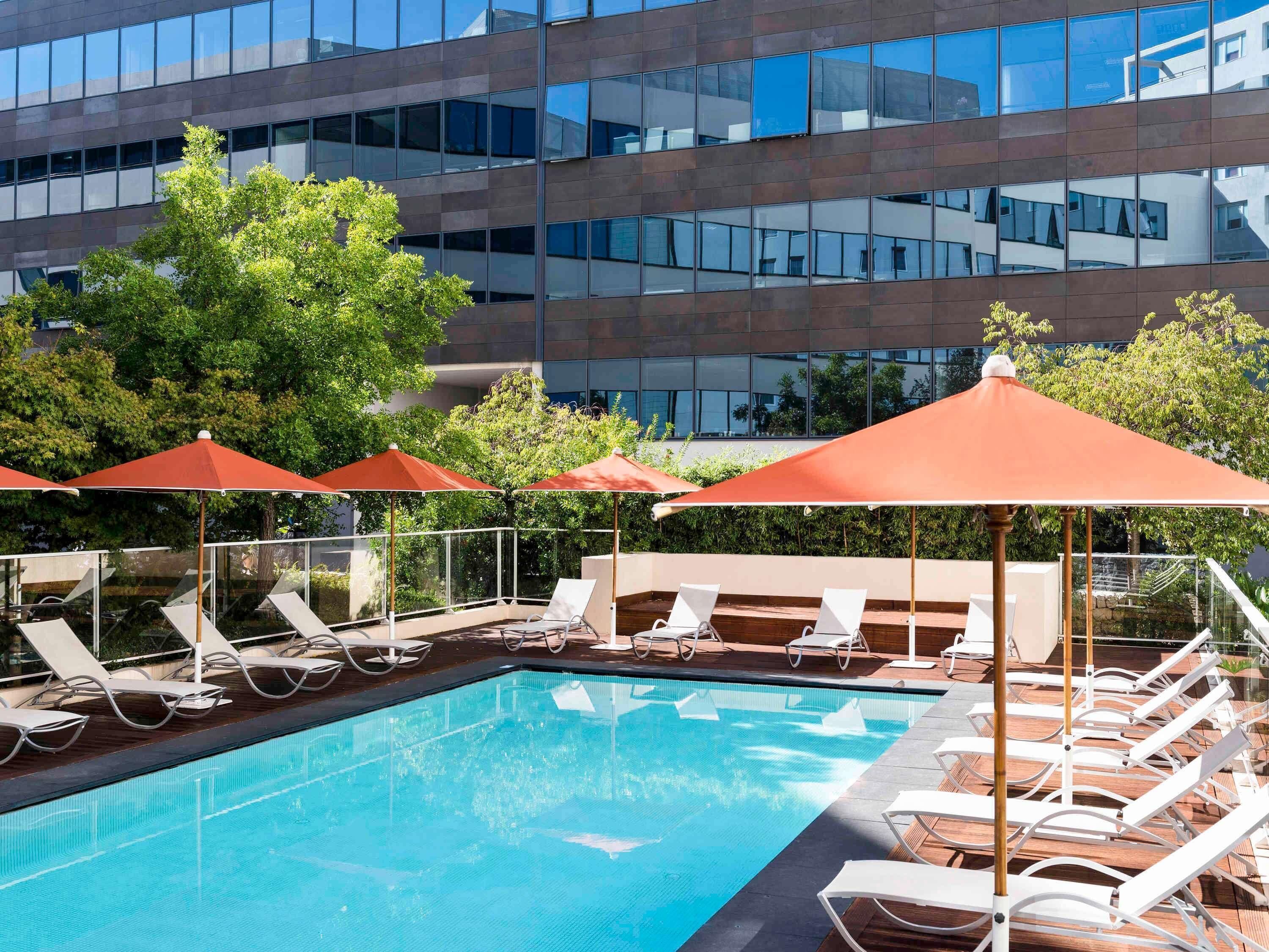 A swimming pool with sun loungers and umbrellas, surrounded by a modern building with reflective glass windows.