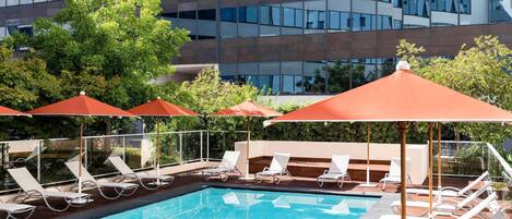 A swimming pool with sun loungers and umbrellas, surrounded by a modern building with reflective glass windows.