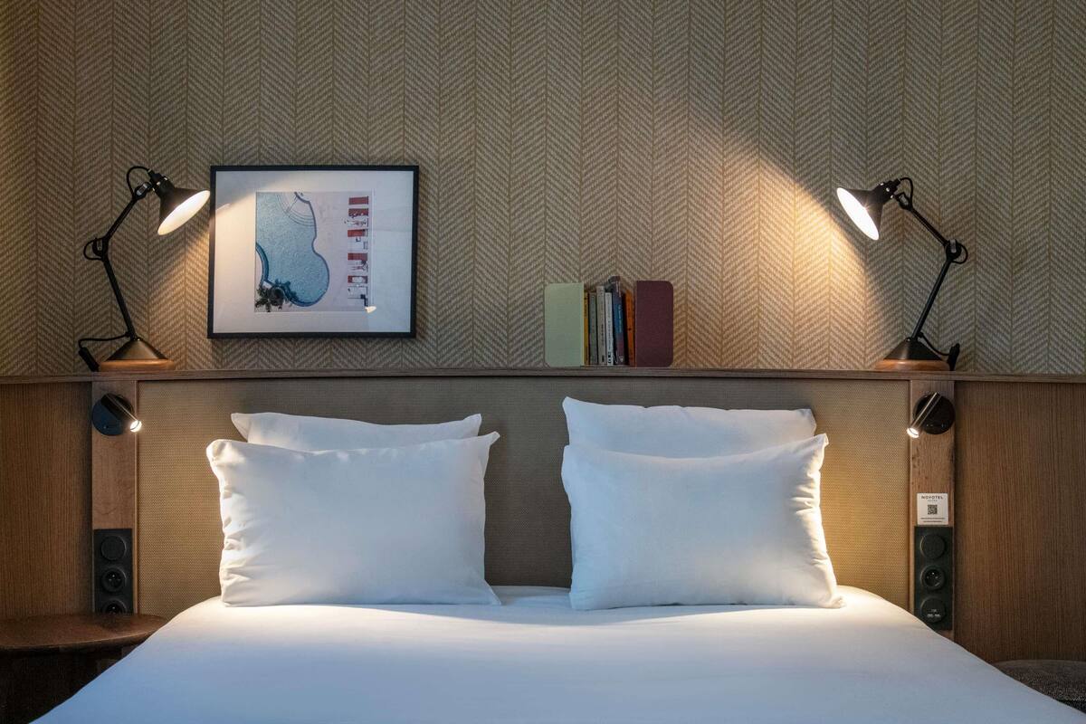 A neatly made bed with two white pillows, a framed artwork above, and two bedside lamps on either side.