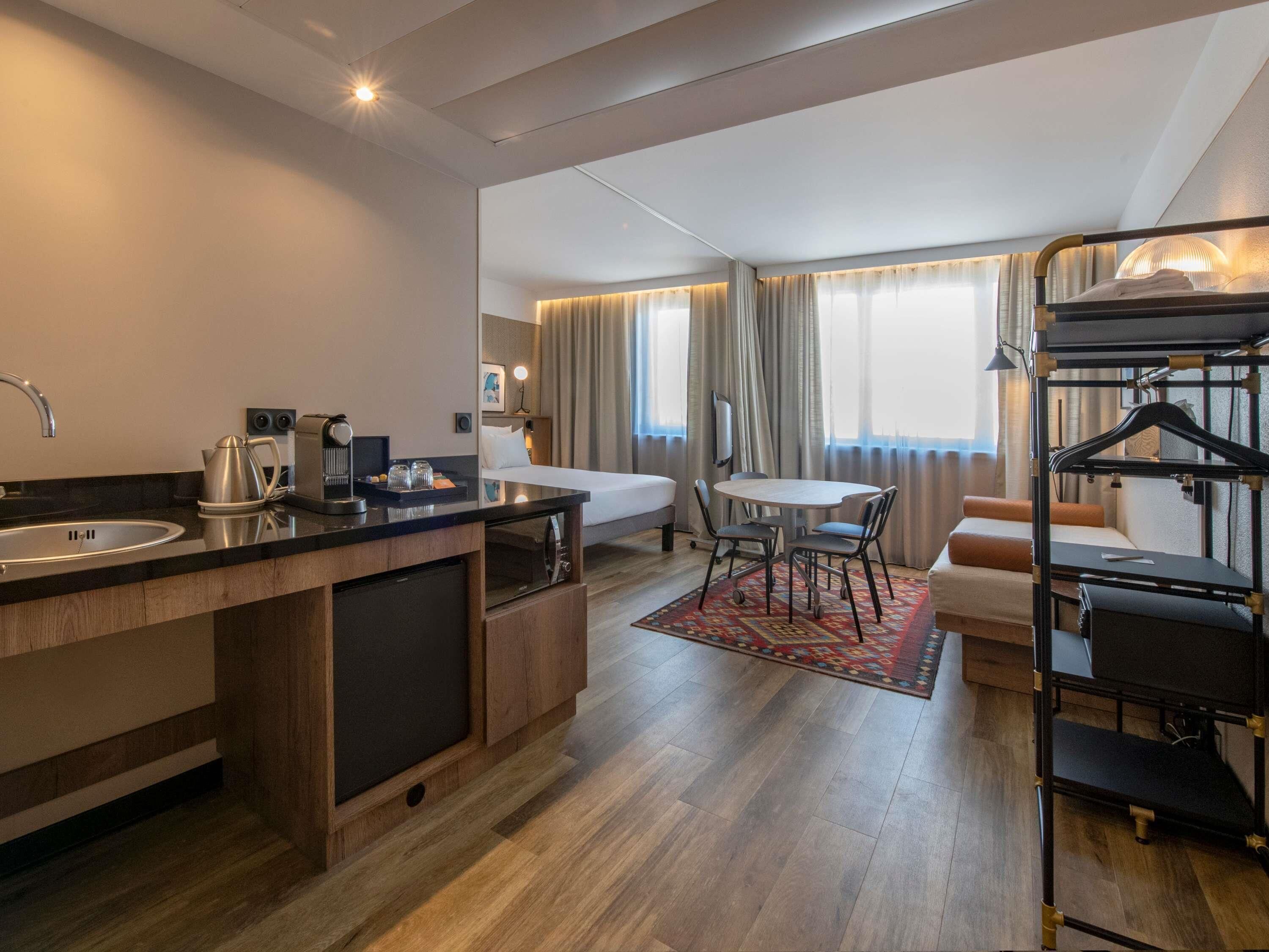 A modern hotel room with a kitchenette, a bed, a dining area, and a bunk bed.
