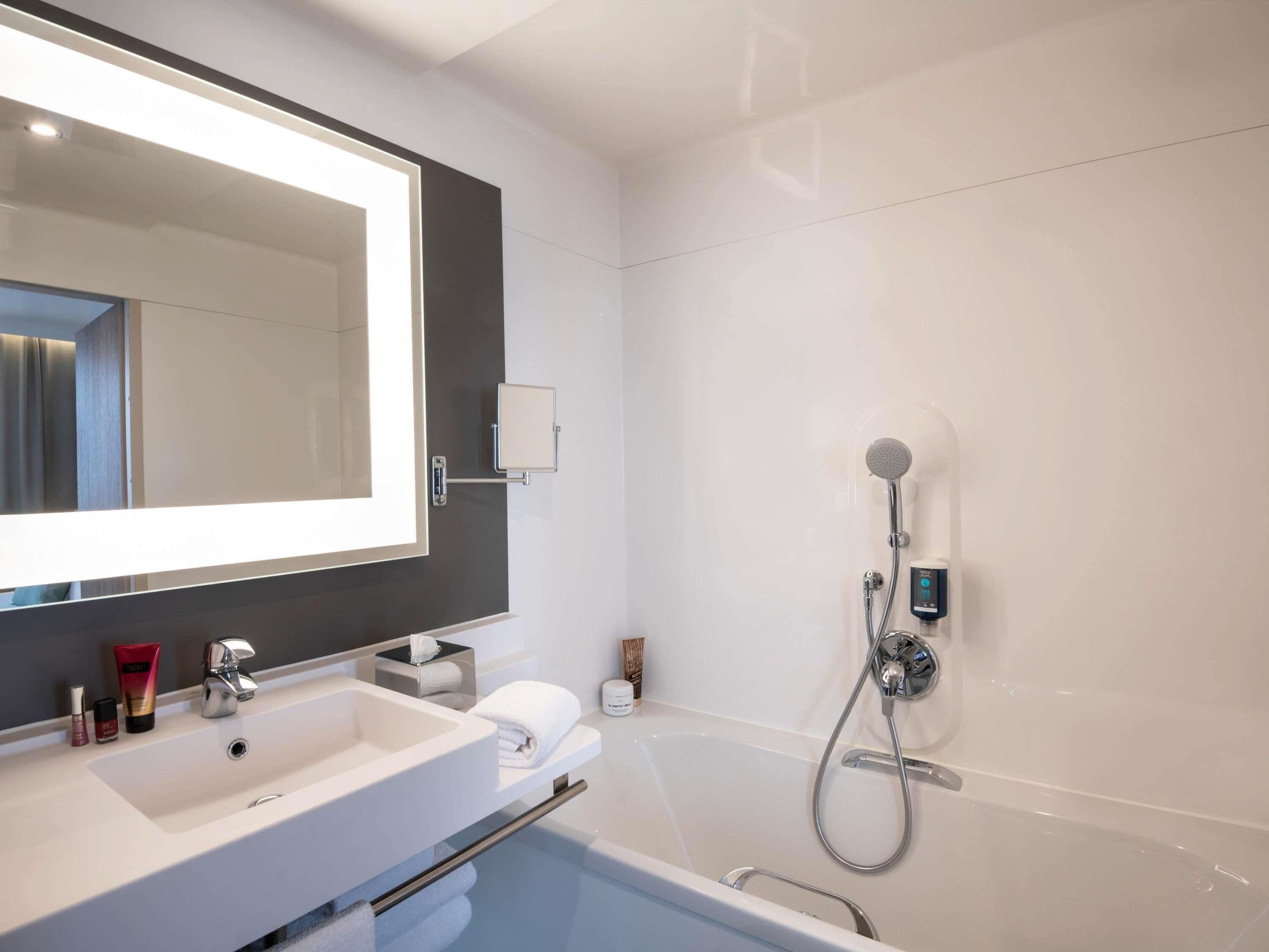 A modern bathroom with a large mirror, a white sink, a handheld showerhead, and a built-in bathtub.