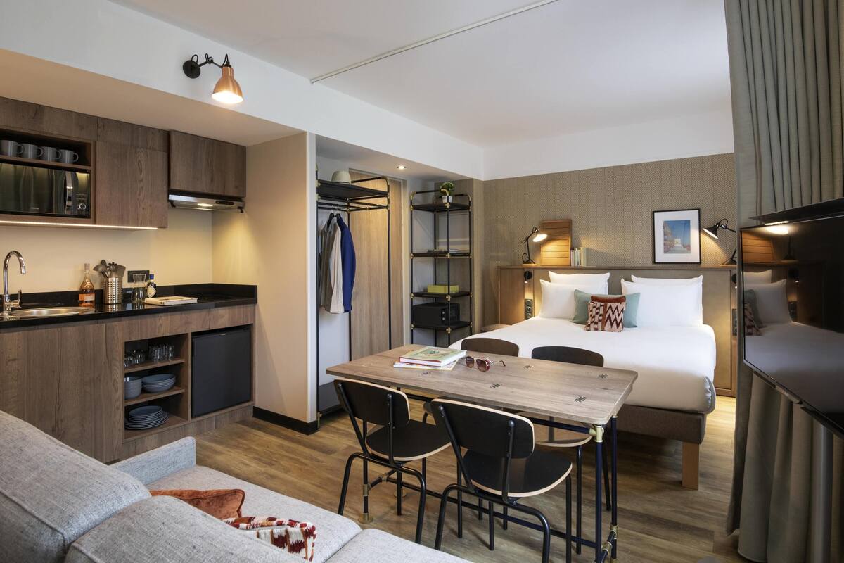 A compact hotel room with a kitchenette, a bed with pillows, a dining table with chairs, and a sofa.