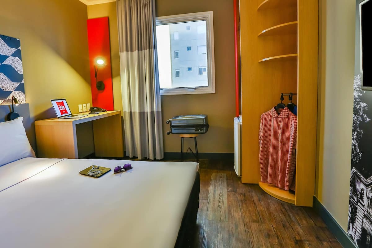 Een hotelkamer met een bed, een bureau met een laptop, een raam met gordijnen, een kapstok en een kledingkast.