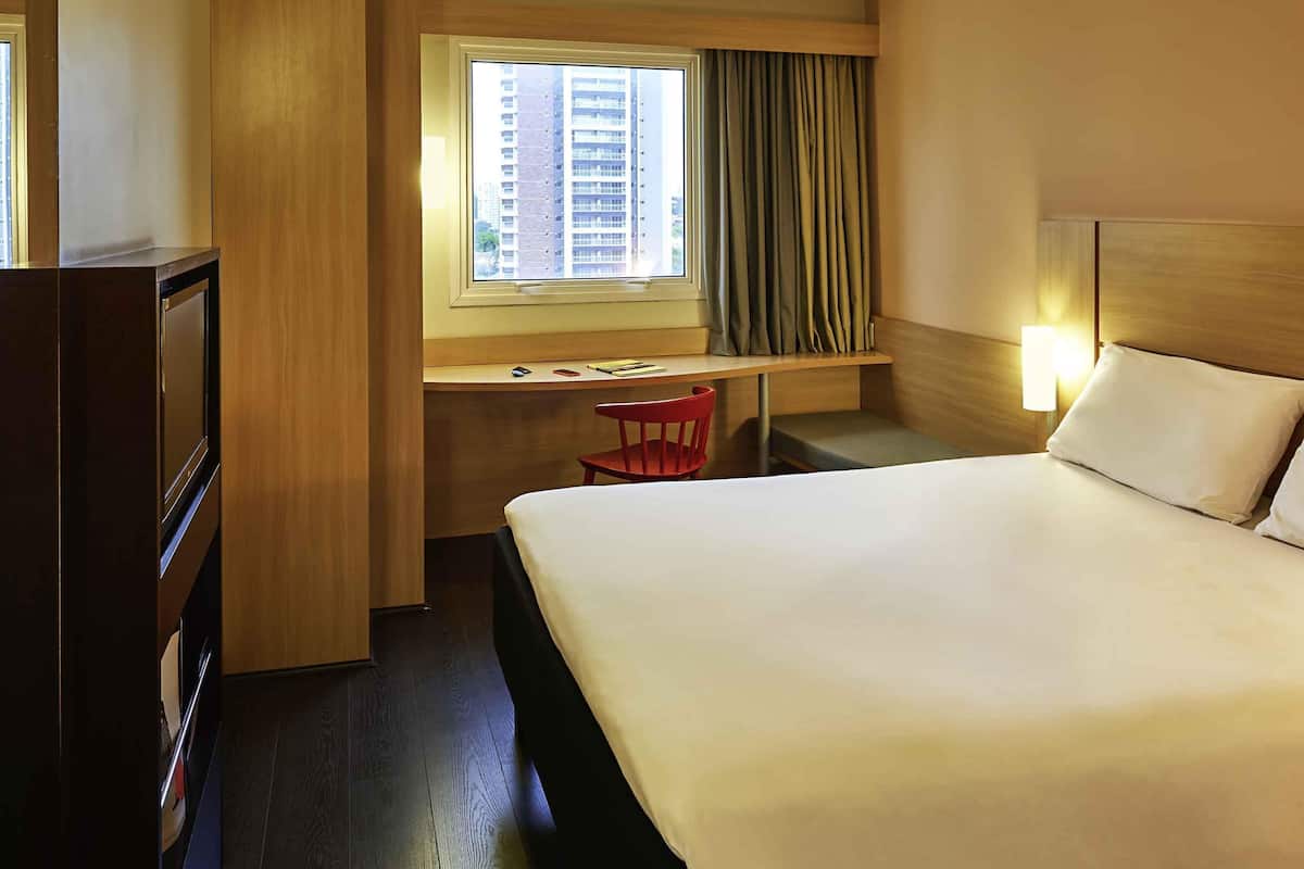 Een hotelkamer met een groot bed, een bureau met stoel, een raam met gordijnen en een wandlamp.