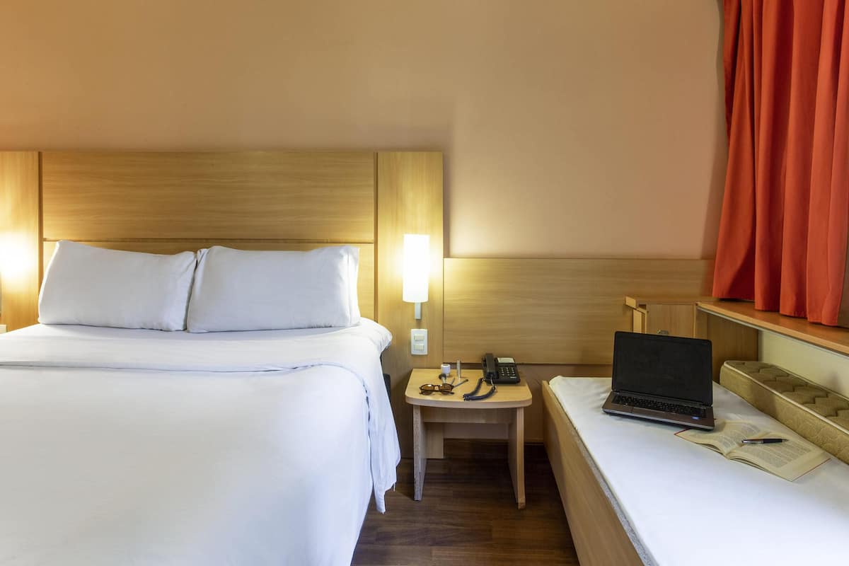 Een hotelkamer met een bed, nachtkastje met een lamp, een laptop en een klein tafeltje met een boek en pen.