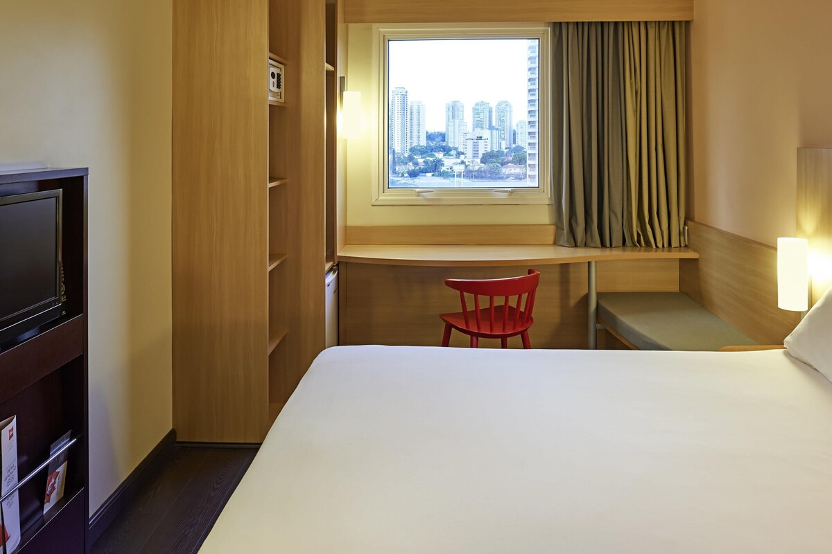 Hotelkamer met een bed, een bureau, een stoel, een televisie en een raam met uitzicht op de stad.