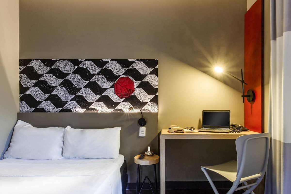 Quarto de hotel com uma cama, uma escrivaninha com um laptop, uma cadeira e uma obra de arte na parede.