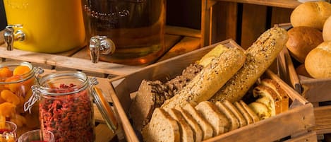 A wooden crate filled with various breads and pastries, a glass jar of dried fruits, and a coffee station with mugs and a kettle.