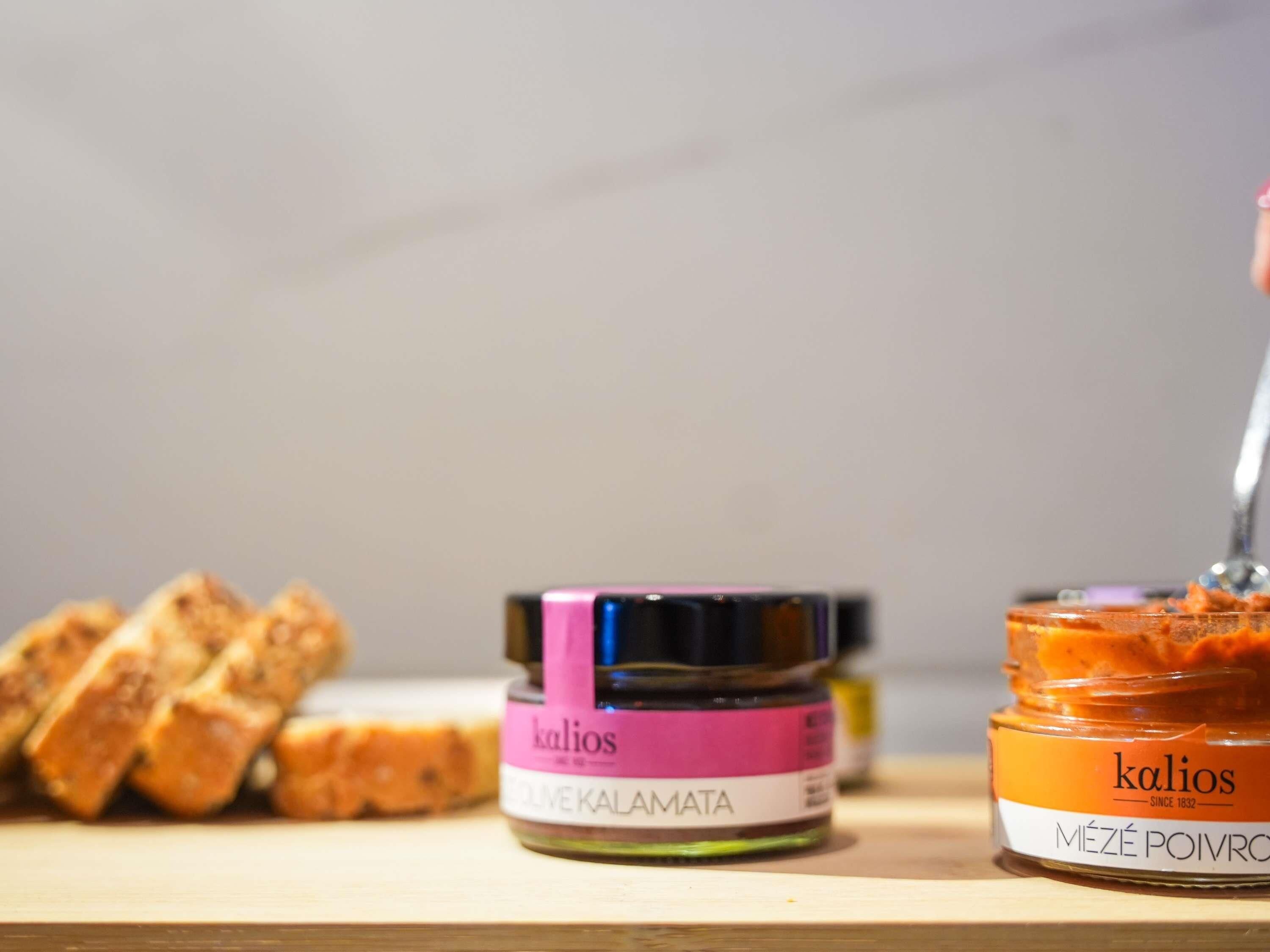 Two jars of Kalios brand products, one labeled 'MÉZÉ POIVRO' and the other 'OLIVEKALAMATA,' alongside some sliced bread on a wooden surface.