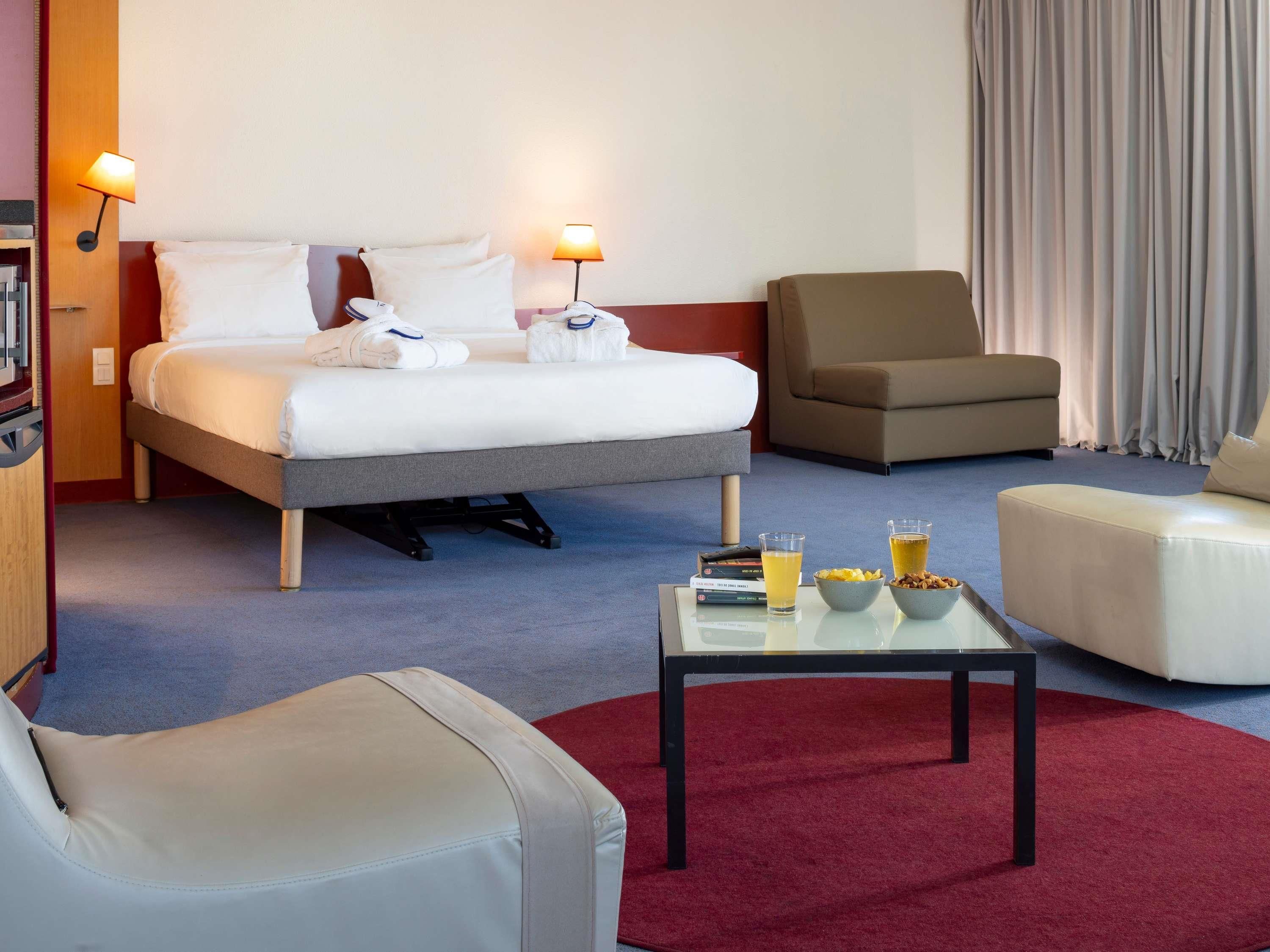 A hotel room with a bed, two bedside lamps, a sofa, a chair, a coffee table with books and glasses, and a red carpet.