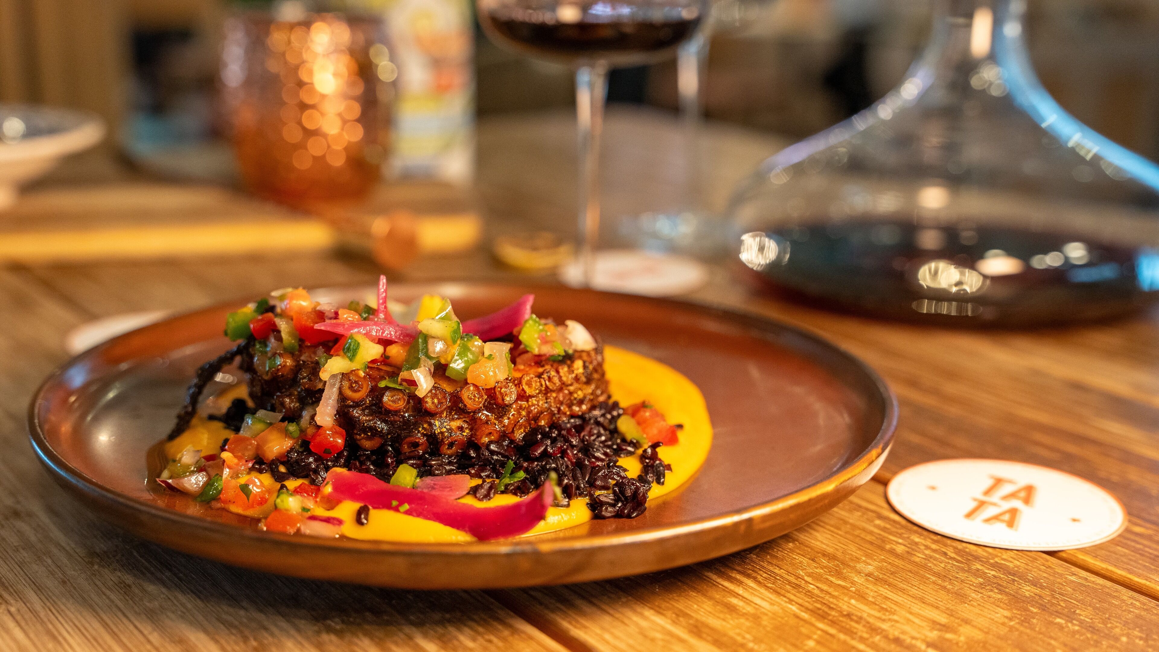 A dish with a variety of colorful toppings on a plate, accompanied by a glass of red wine and a decorative bottle on a wooden table.