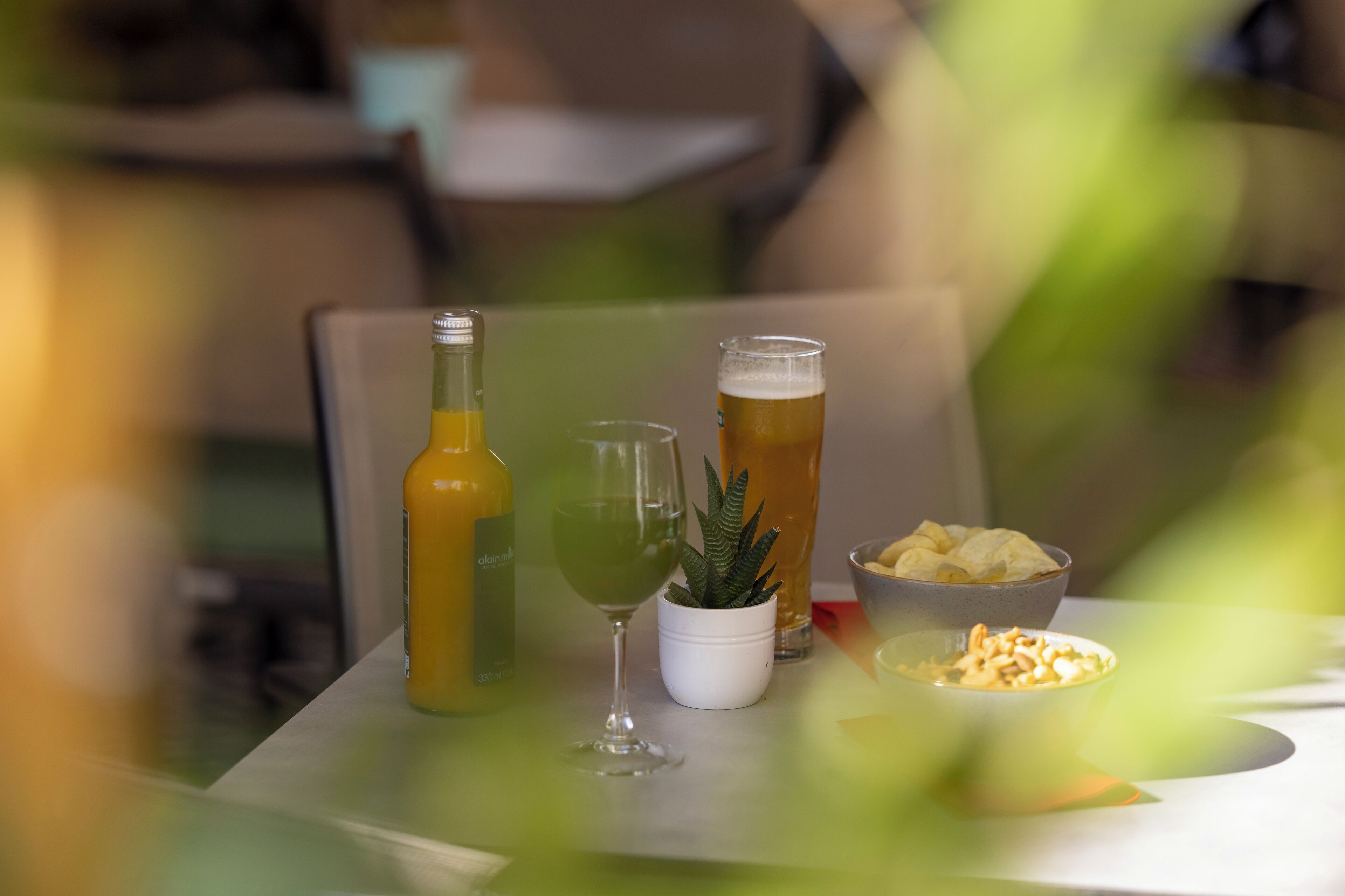 Uma mesa com uma garrafa, um copo, um vaso de planta e recipientes com petiscos.