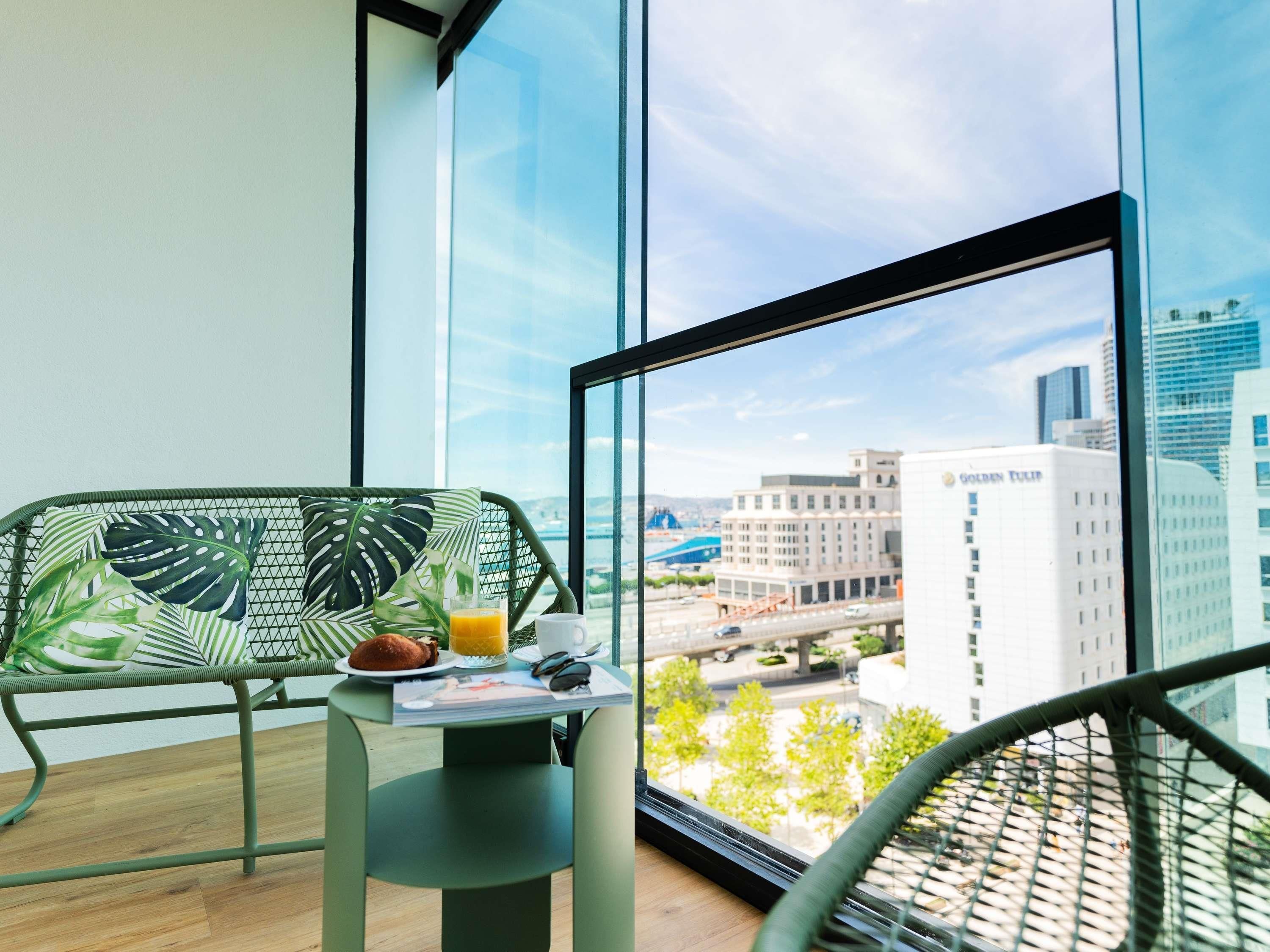 Un balcón con vistas a un paisaje urbano, una silla verde, una mesita con una bebida y un plato de comida, y una ventana con marco negro.