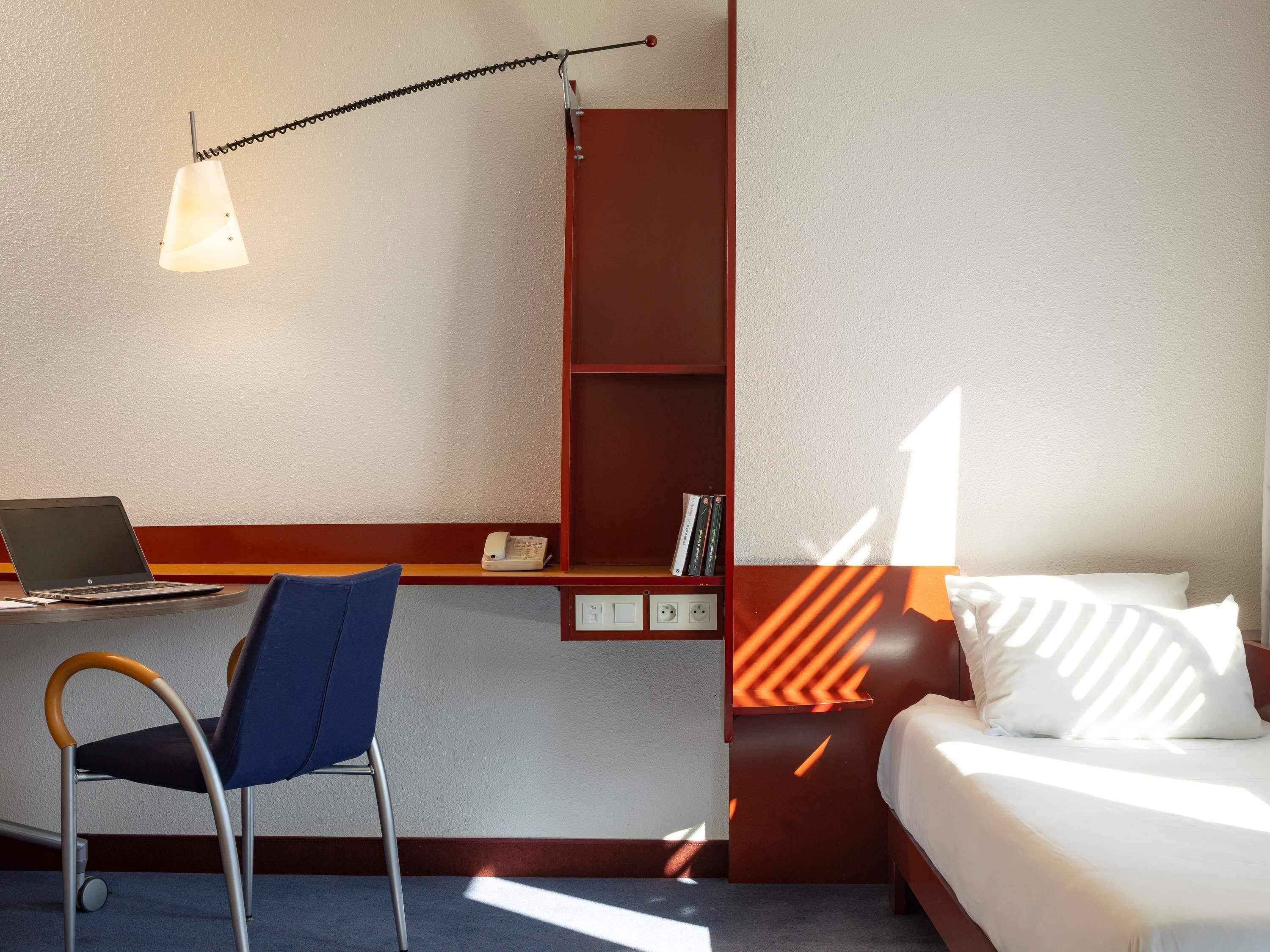 A hotel room with a desk, chair, bed, and a bookshelf.