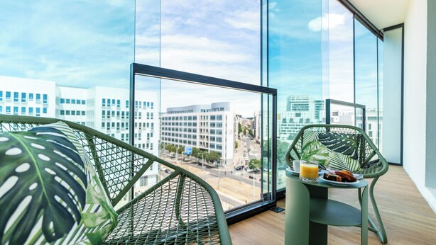 Un balcón con vistas a edificios de la ciudad, una mesa con alimentos para el desayuno y sillas verdes.