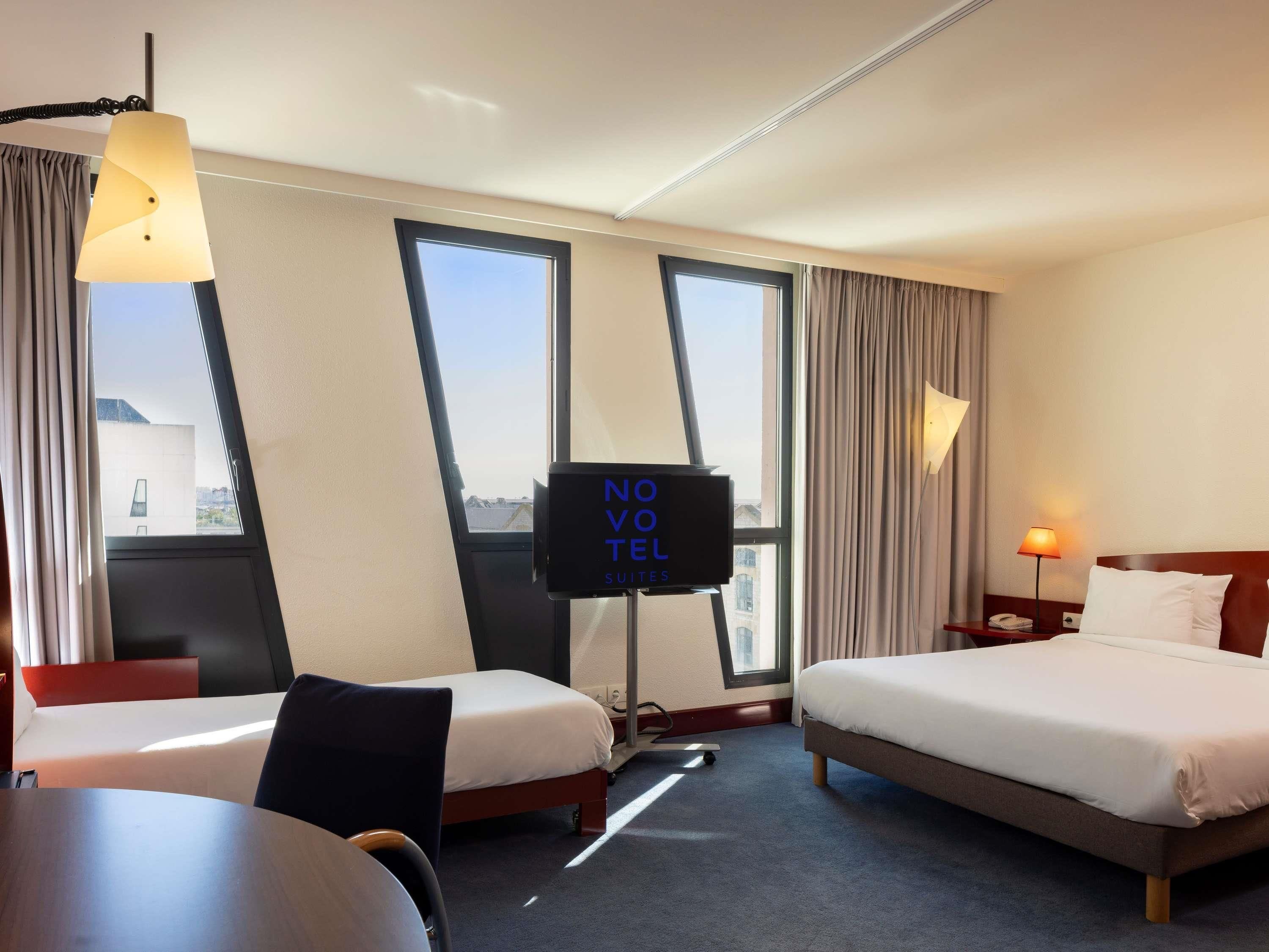 A hotel room with two beds, a desk, a TV, and large windows.