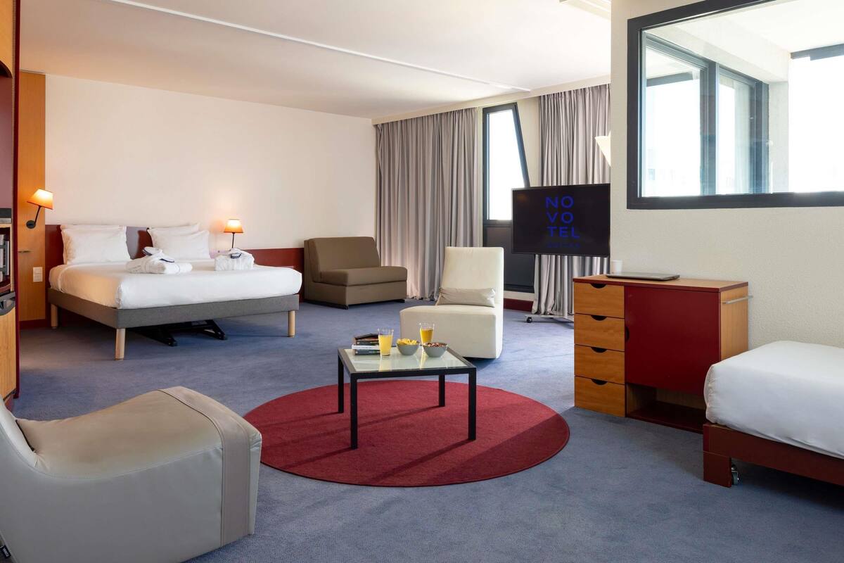 Ein Hotelzimmer mit Bett, zwei Sesseln, einem Schreibtisch, einem Fernseher und einem fenster mit Vorhängen.