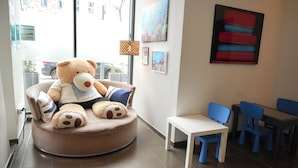 Una habitación con un gran osito de peluche en un sillón redondo, una mesita, sillas azules y obras de arte en las paredes.