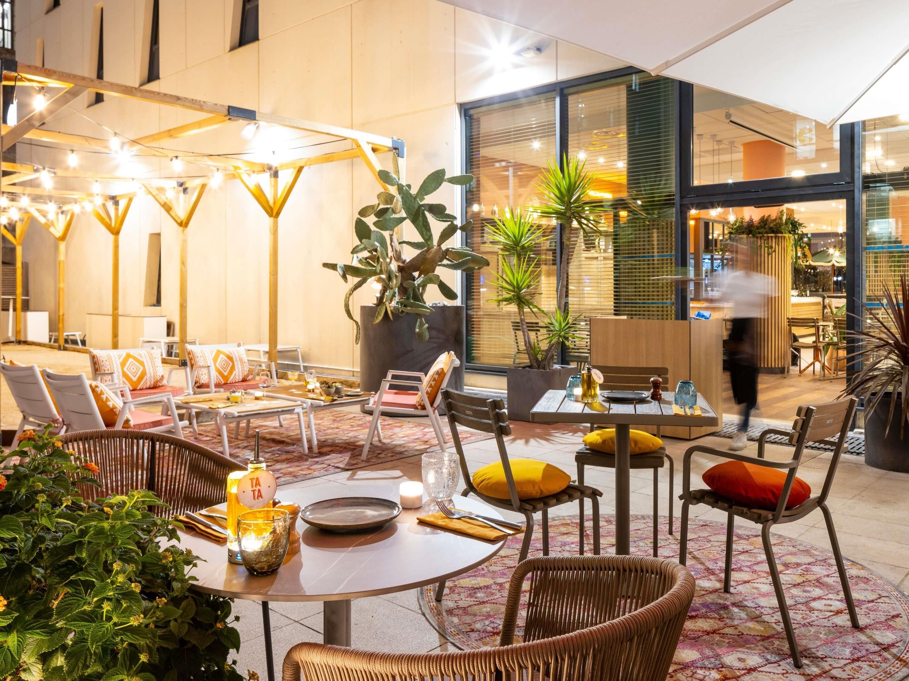 Un espace de détente intérieur moderne, doté de tables rondes et de chaises en osier, agrémenté de plantes en pot et d’un éclairage d’ambiance.