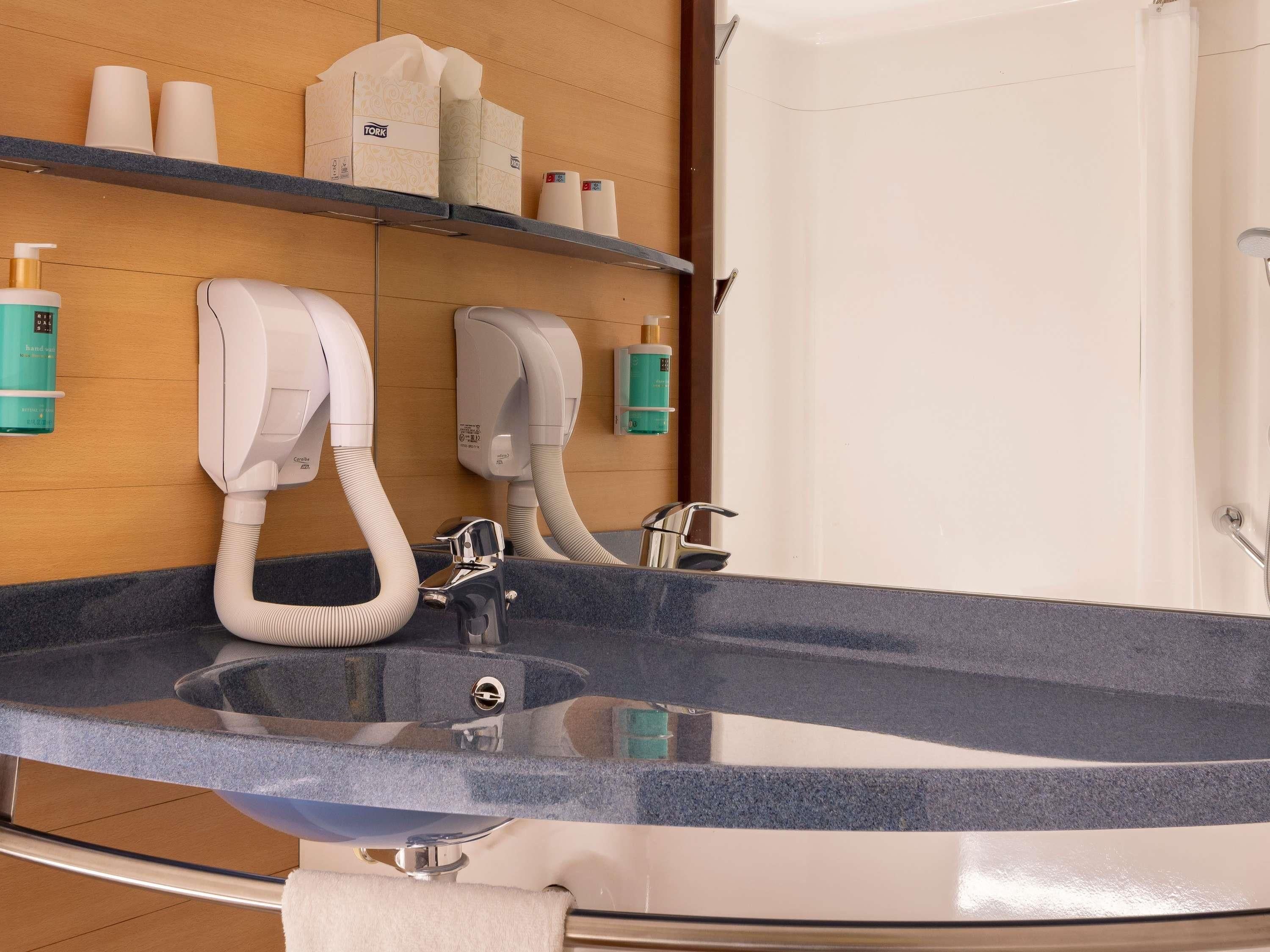 Ein modernes Badezimmer mit einem großen Waschbecken, zwei Handtrocknern und Regalen mit Toilettenartikeln.