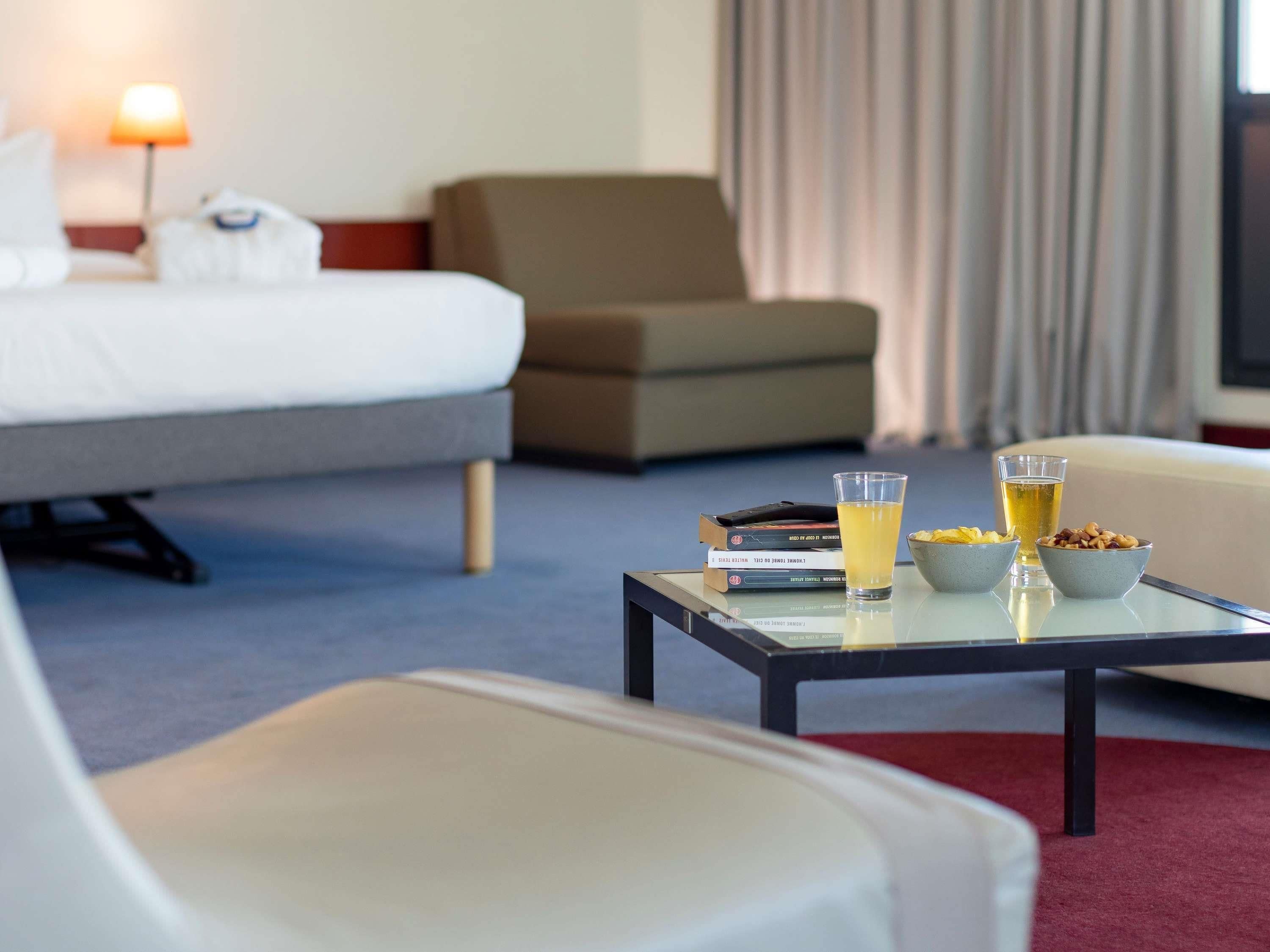 A hotel room with a bed, a sofa, a coffee table with books and drinks, and a lamp.