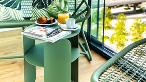 Una silla verde de mimbre con una revista, una taza, un plato con comida y un vaso de jugo de naranja sobre una mesita.