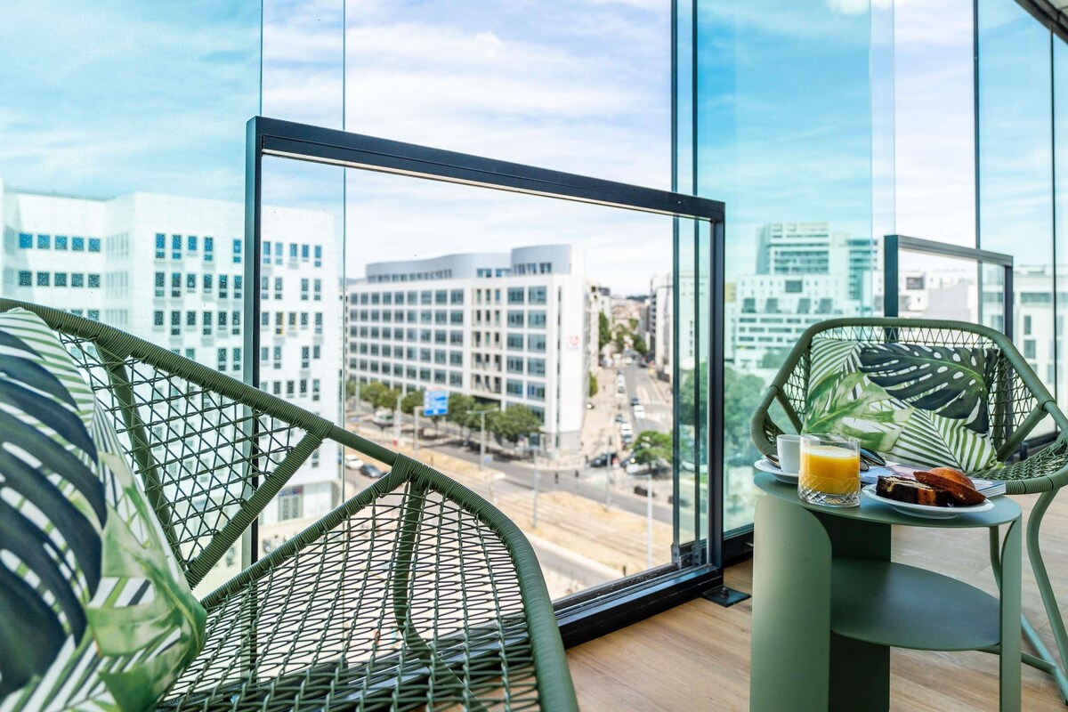 A balcony with a city view, a wicker chair, a small table with a glass of orange juice and a plate of food.
